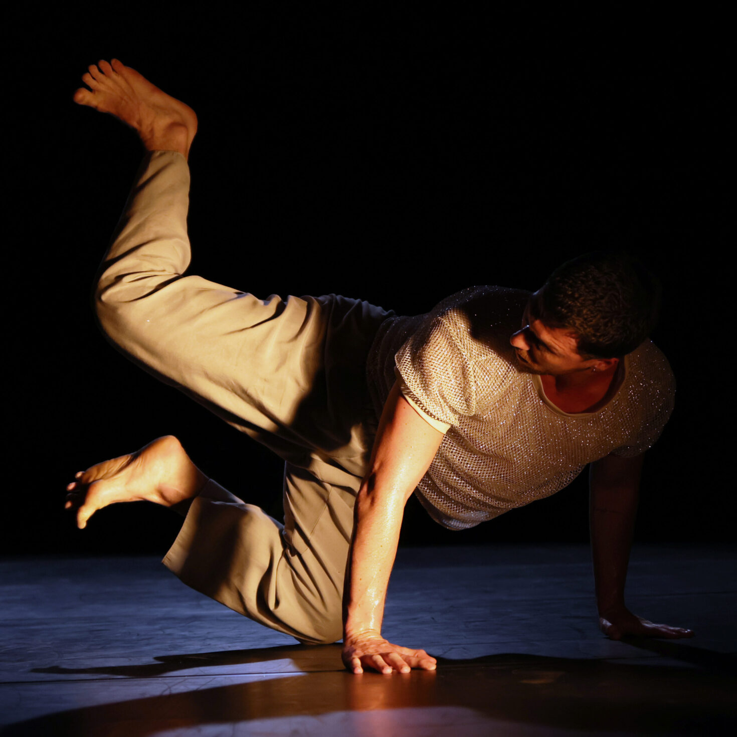 Irene K. - Auf der Bühne der TanzArt OstWest 2025 führt eine Person eine zeitgenössische Tanzbewegung aus und balanciert dabei anmutig im schimmernden Licht, wobei ihre weite Hose und ihr funkelndes Oberteil vor dem dunklen Hintergrund des Alten Schlachthofs in Eupen besonders zur Geltung kommen.