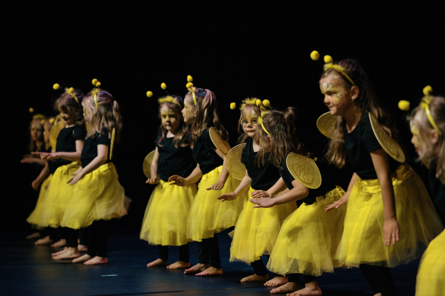 Irene K. - Eine Gruppe kleiner Kinder, die als Bienen verkleidet sind – mit gelben Tutus, Flügeln und Stirnbändern –, tritt barfuß auf der Bühne auf und präsentiert vor einem dunklen Hintergrund Tanzbewegungen, die vom Hip-Hop und vom zeitgenössischen Tanz inspiriert sind.