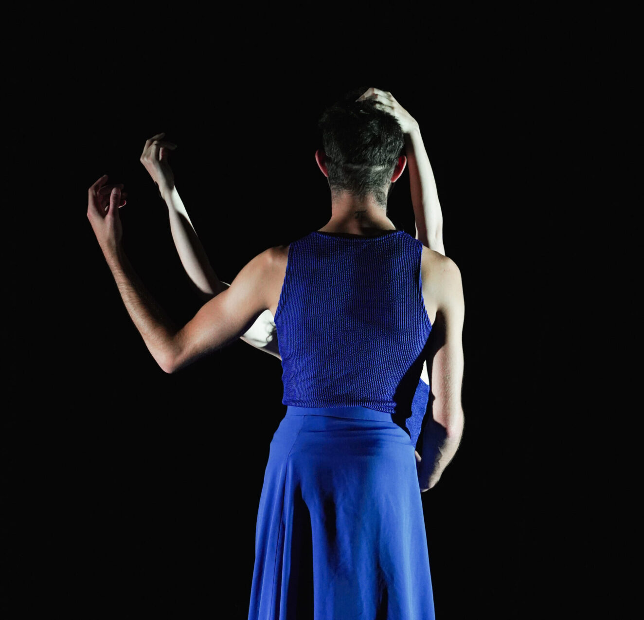Irene K. - A person in a blue sleeveless top and skirt stands with their back to the camera, arms raised, as another’s pale arms intertwine with theirs. Both are highlighted against a dark background, capturing the essence of Tanzperformance and Bühnenkunst.