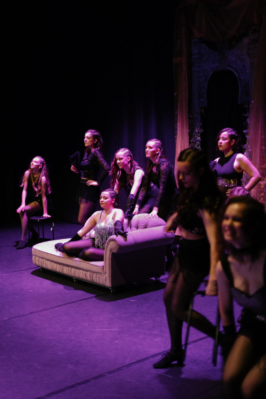 Irene K. - Eine Gruppe von Darstellern in schwarzen Kostümen sitzt und steht auf der Bühne um ein beiges Sofa herum, das während einer Tanzaufführung von dramatischem violettem Licht beleuchtet wird, während im Hintergrund ein dunkler Vorhang und ein verzierter Torbogen zu sehen sind.