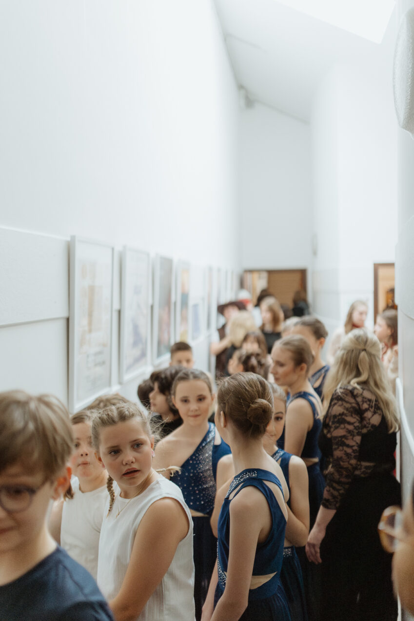 Irene K. - Eine Gruppe von Kindern und Jugendlichen in festlicher Kleidung, darunter blaue und weiße Kleider, steht in einem hell erleuchteten Flur im Tanzstudio Eupen und wartet in einer Reihe auf eine Veranstaltung oder Aufführung von „Kreativer Tanz“, umgeben von gerahmten Bildern.