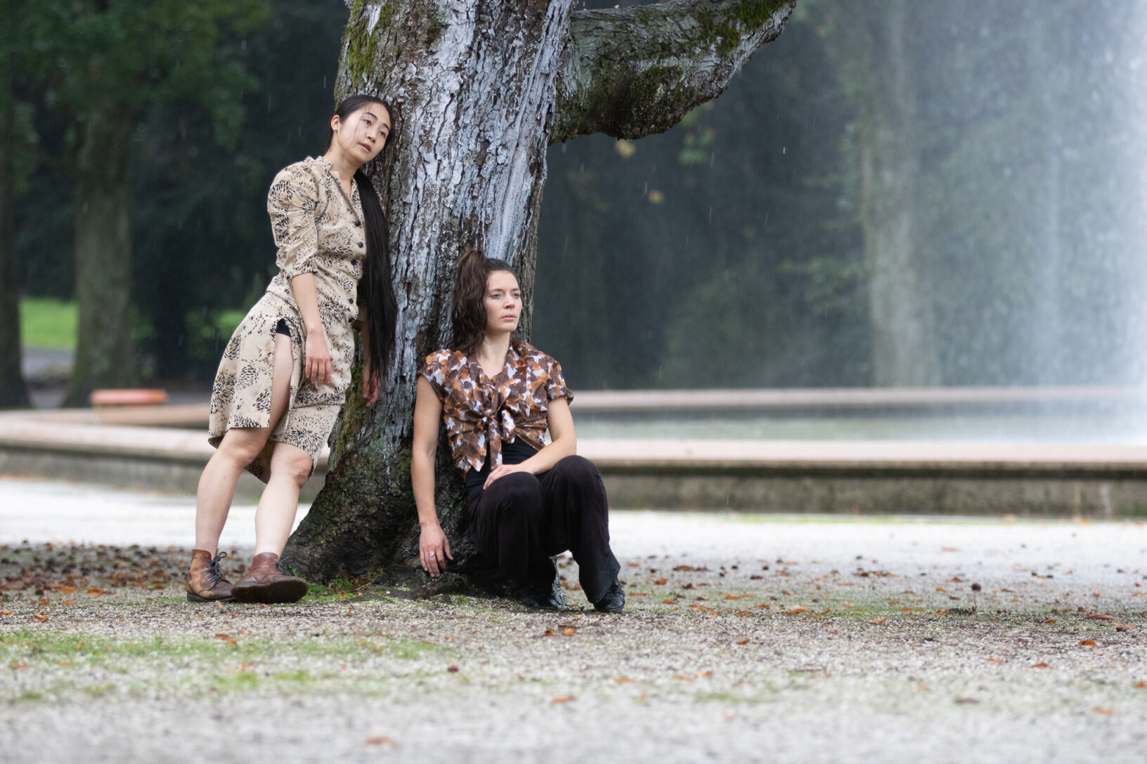 Irene K. - Zwei Frauen posieren an einem großen Baum; die eine steht und lehnt sich an den Stamm, während die andere am Fuß des Baumes kauert, was an zeitgenössischen Tanz erinnert. Im Hintergrund sind ein Wasserbrunnen und Grünpflanzen zu sehen.