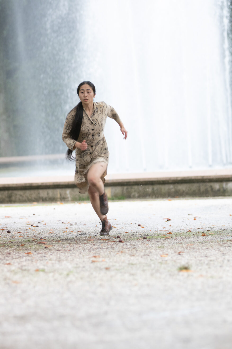Irene K. - Eine Frau mit langen dunklen Haaren, die ein gemustertes Kleid trägt und zeitgenössischen Tanz verkörpert, läuft auf einem Kiesweg mit einem großen Wasserbrunnen im Hintergrund auf die Kamera zu.