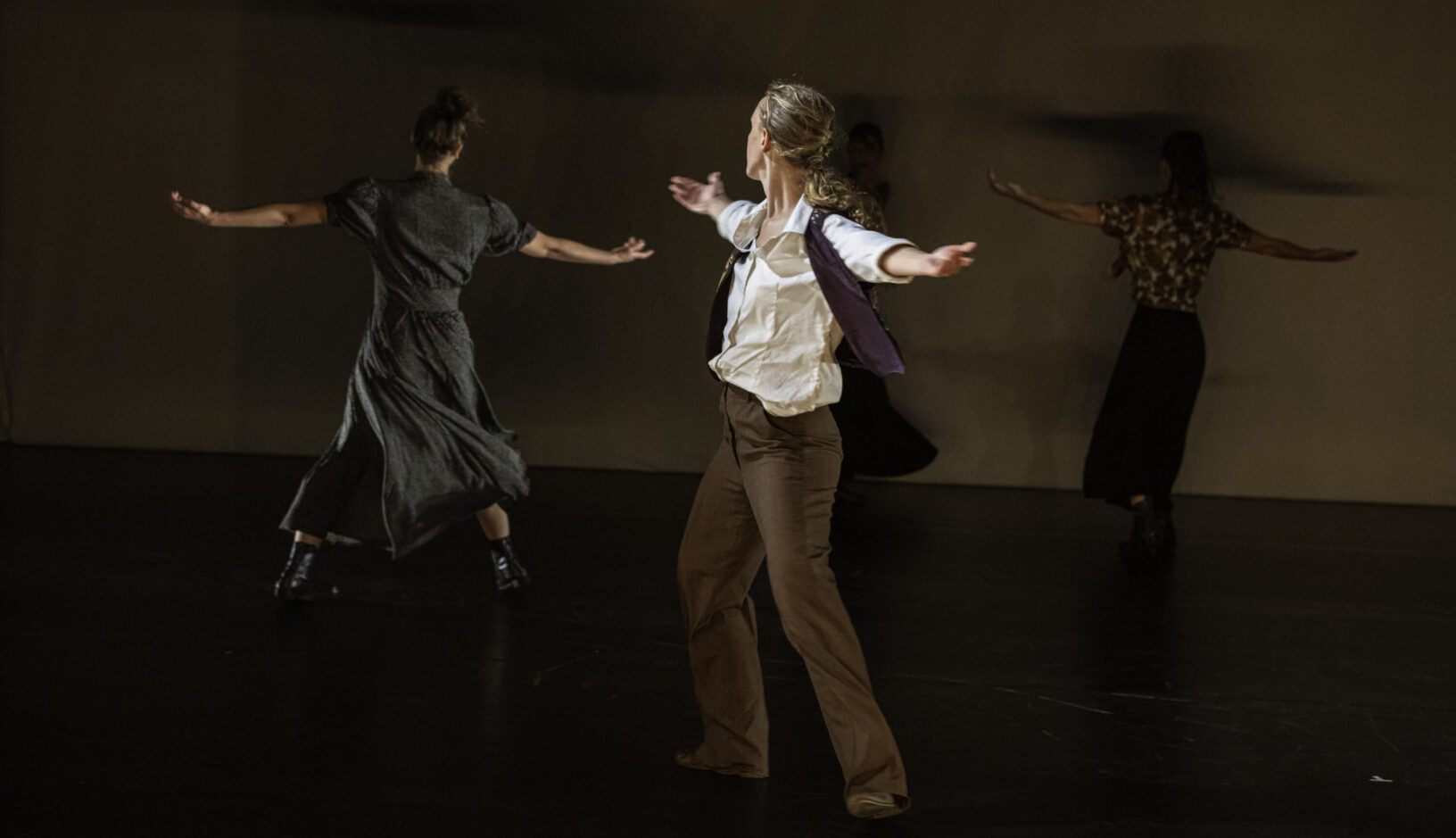 Irene K. - Vier Personen tanzen auf einer schwach beleuchteten Bühne, jede mit ausgestreckten Armen und in weitem Abstand, ihre dunkle Freizeitkleidung verschmilzt mit dem schlichten Hintergrund. Die Choreografie der Compagnie Irene K. unterstreicht jede Bewegung in diesem eindrucksvollen Beispiel der Bühnenkunst.