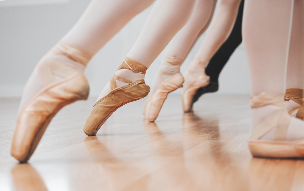 Irene K. - Nahaufnahme der Beine und Füße von Balletttänzern in Spitzenschuhen, die auf einem hölzernen Studioboden auf der Spitze stehen. Die Tänzerinnen und Tänzer stehen in einer Reihe nebeneinander, ihre Balance und Anmut spiegeln den Geist der Modern Jazz Tanzkurse in Verviers wider.