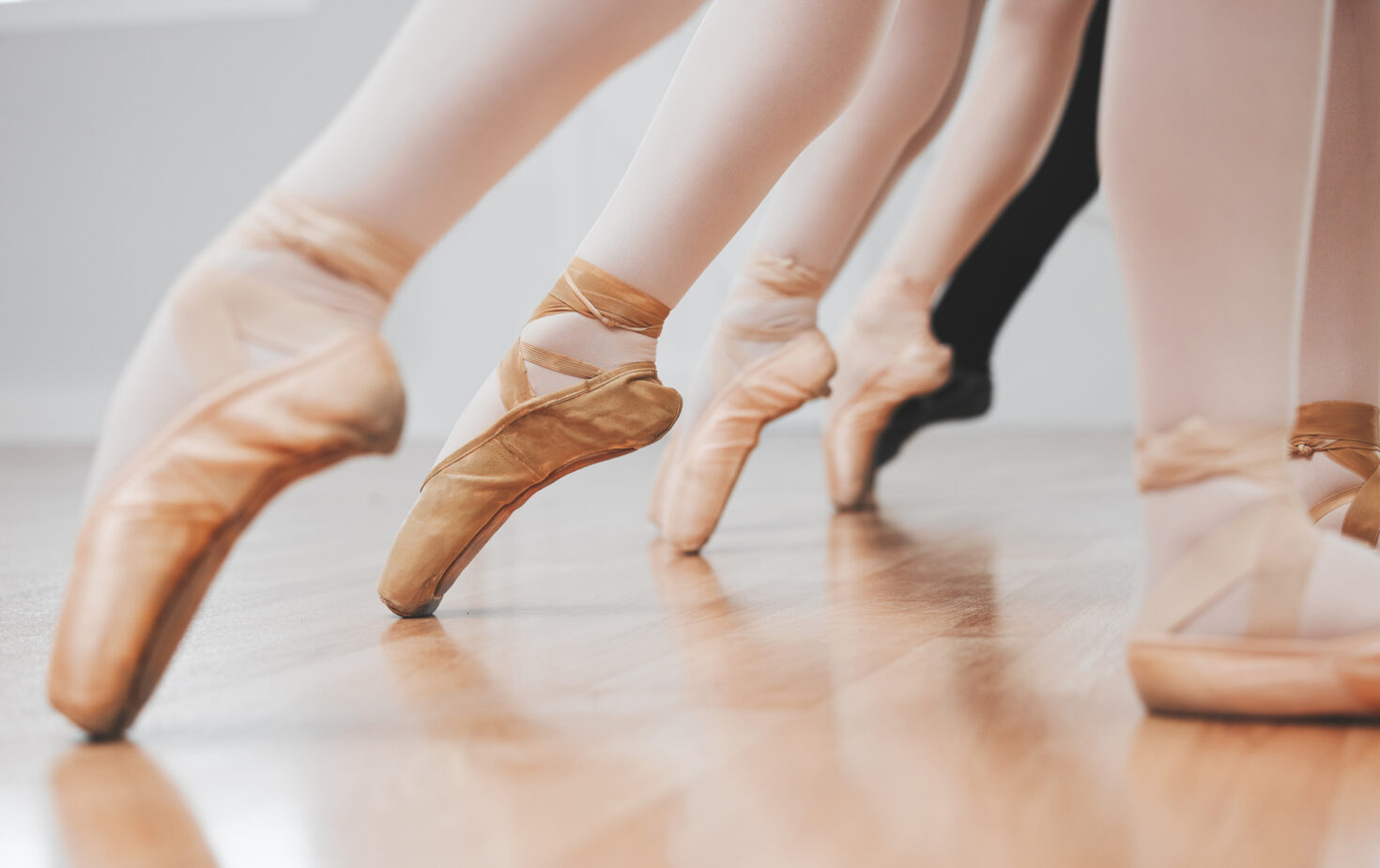 Irene K. - Nahaufnahme der Beine und Füße von Balletttänzern in Spitzenschuhen, die auf einem hölzernen Studioboden auf der Spitze stehen. Die Tänzerinnen und Tänzer stehen in einer Reihe nebeneinander, ihre Balance und Anmut spiegeln den Geist der Modern Jazz Tanzkurse in Verviers wider.