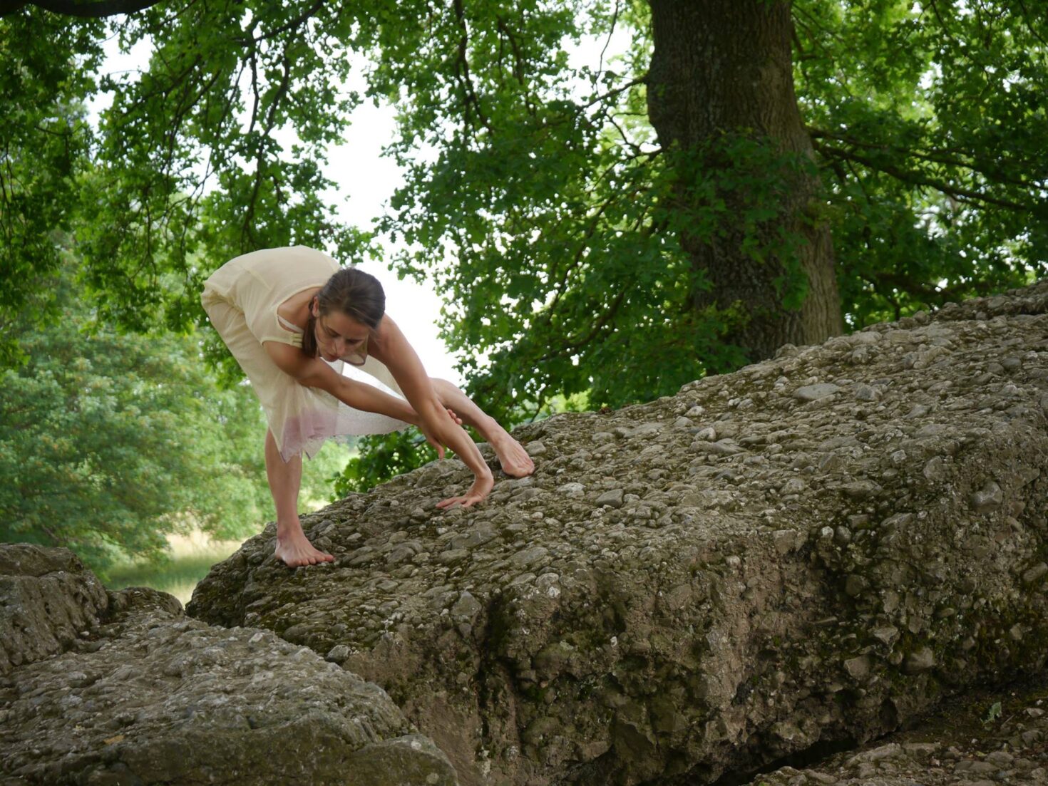 Irene K. - Eine Frau in einem leichten Kleid balanciert anmutig auf einem großen, unebenen Felsen im Freien, umgeben von grünen Bäumen - ihre Pose erinnert an die Fluidität des zeitgenössischen Tanzes und die Ausdruckskraft der Tanzperformance.