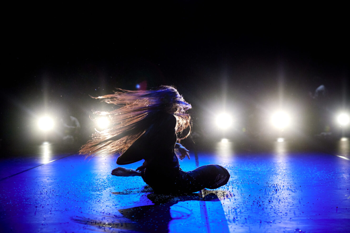 Irene K. - A person with long hair sits on a stage floor, dramatically flipping their hair as bright lights shine behind them, casting blue and white highlights—a powerful moment of Bühnenkunst by the professionelle Tanzcompagnie Compagnie Irene K.