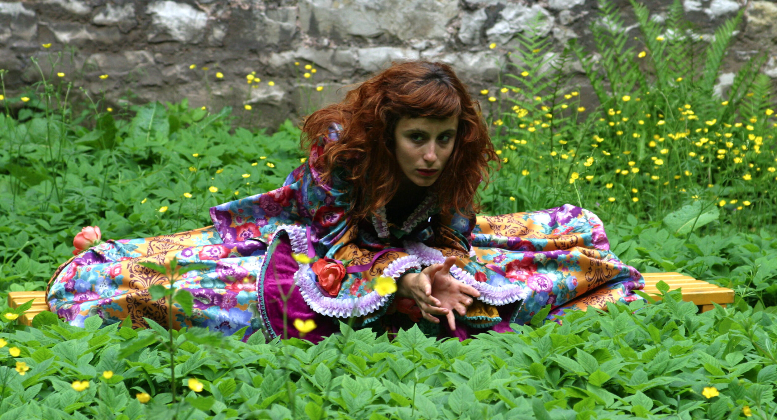 Irene K. - Eine rothaarige Person in einem bunten, gemusterten Outfit sitzt auf einer gelben Bank in einem üppigen Garten, als würde sie zwischen den Bewegungen einer Choreografie innehalten, umgeben von grünen Pflanzen und gelben Blumen, mit einer Steinmauer im Hintergrund.