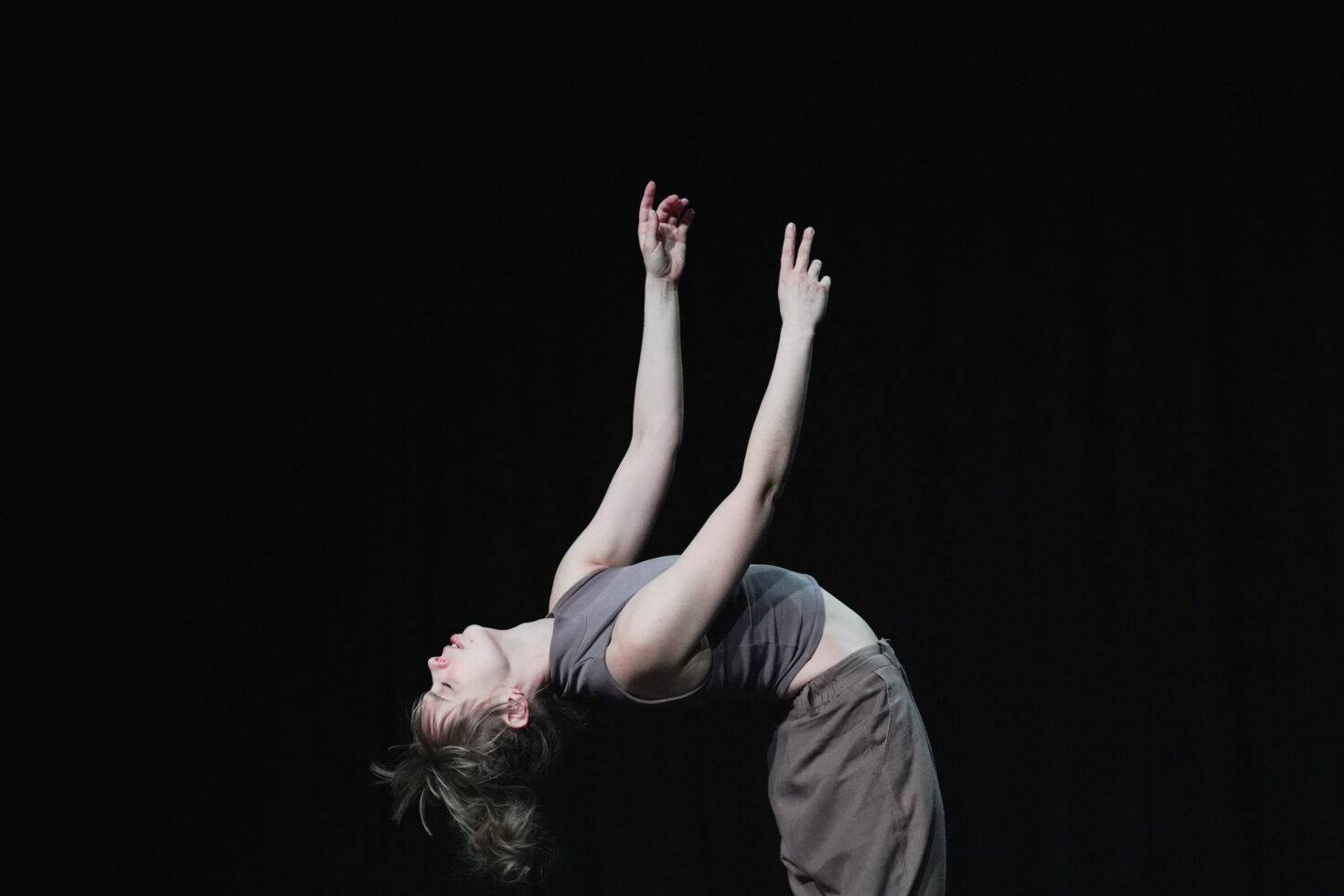 Irene K. - Eine Person mit hellem Haar und einem ärmellosen Oberteil beugt sich mit erhobenen Armen theatralisch nach hinten und bringt so in dieser eindrucksvollen Tanzperformance vor dunklem Hintergrund die Ausdruckskraft der Bühnenkunst zum Ausdruck.