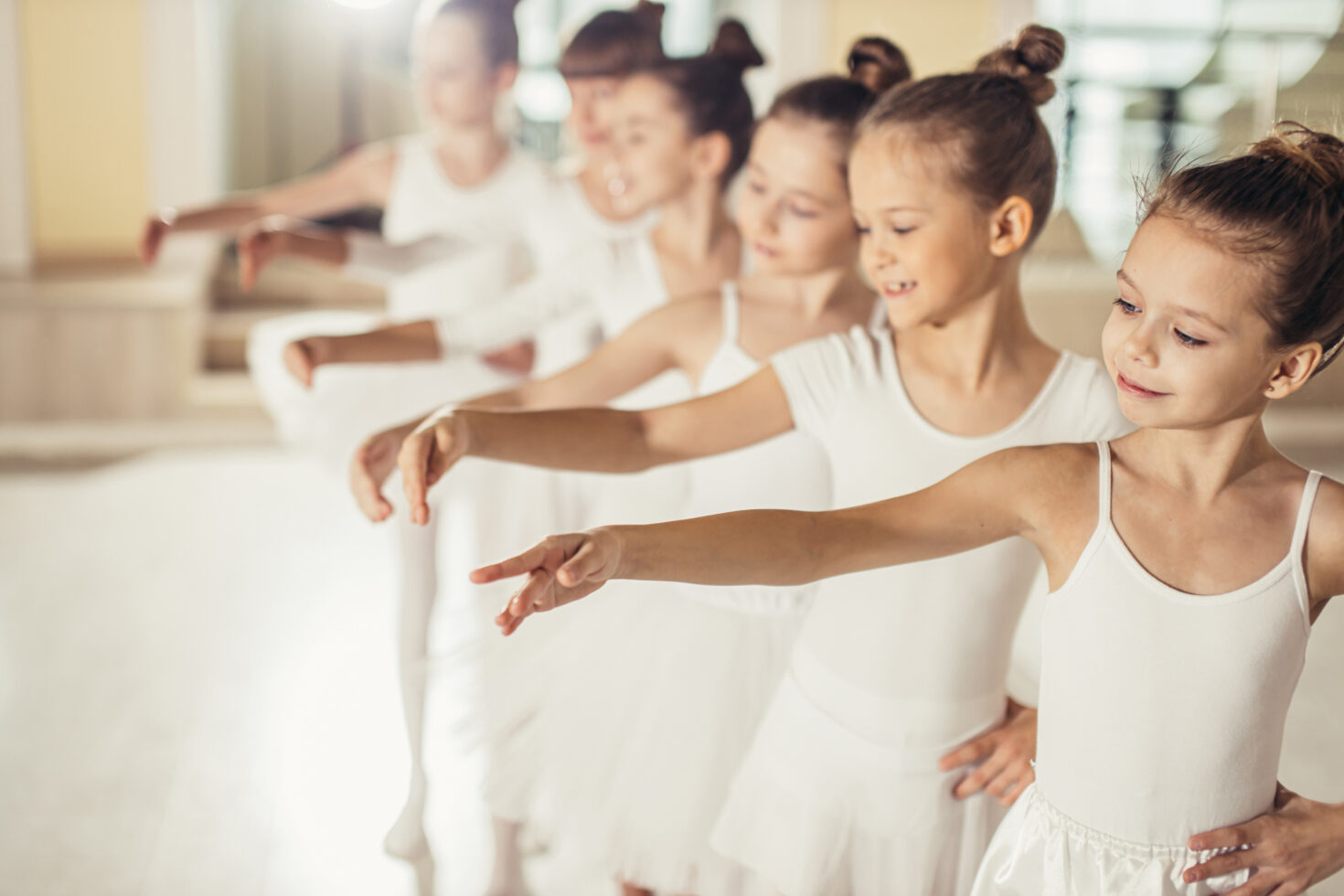 Irene K. - Eine Gruppe junger Mädchen in weißen Ballettkleidern übt an einer Ballettstange. Sie stehen in einer Reihe mit anmutig ausgestreckten Armen, lächeln und konzentrieren sich auf ihre Positionen in einem hell erleuchteten Tanzstudio.