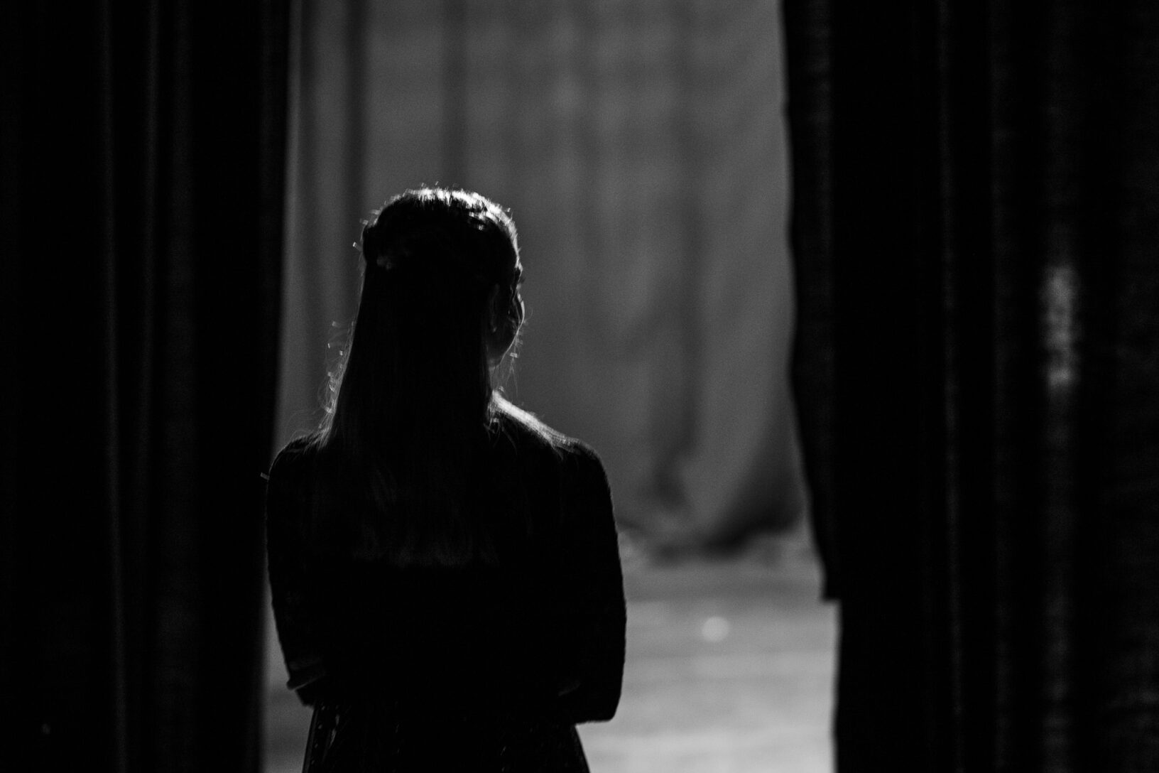 Irene K. - Eine Person steht hinter den Kulissen des Tanzstudios Eupen im Schatten und blickt durch die halb geöffneten Vorhänge auf die hell erleuchtete Bühne, wobei sie der Kamera den Rücken zukehrt – in einer Schwarz-Weiß-Kulisse.