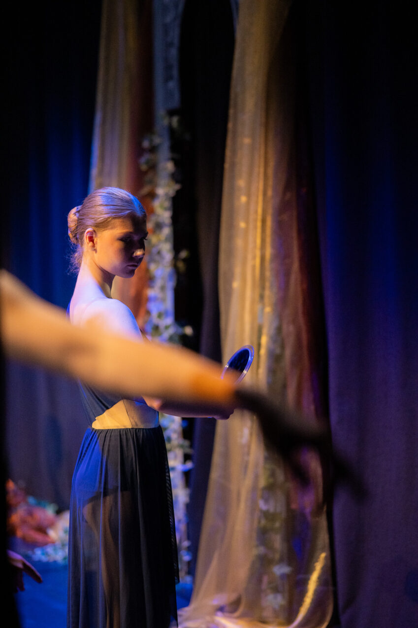 Irene K. - Eine Balletttänzerin in einem dunklen, eleganten Kostüm steht auf der Bühne und hält einen runden Gegenstand in der Hand, während im Hintergrund wallende Vorhänge und sanfte Beleuchtung zu sehen sind. Im Vordergrund streckt eine weitere Tänzerin den Arm aus und vermittelt so die Anmut des kreativen Tanzes.