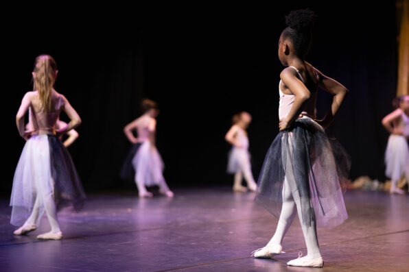 Irene K. - Junge Balletttänzerinnen in Tutus stehen auf einer Bühne und üben mit den Händen in den Hüften. Der Fokus liegt auf einem Mädchen, während die anderen im Hintergrund verschwimmen. Warme, theatralische Beleuchtung sorgt für die richtige Stimmung – perfekt für vielfältige Tanzkurse wie zeitgenössischen Tanz oder Ballett.