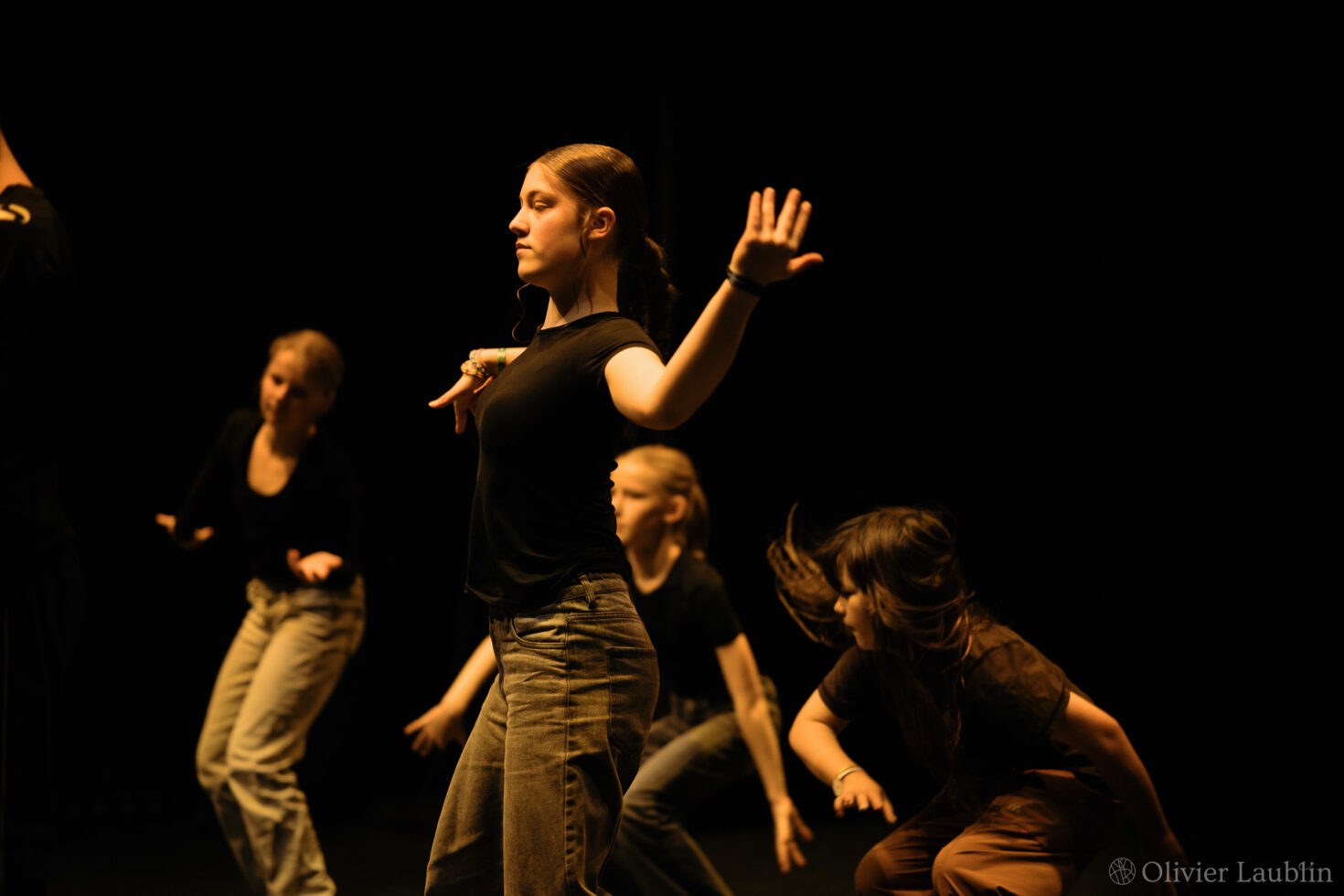Irene K. - Eine Gruppe junger Menschen in Freizeitkleidung führt auf einer schwach beleuchteten Bühne in einer Tanzschule in Eupen eine dynamische Tanzchoreografie auf, wobei dramatische Schatten und warmes Licht ihre ausdrucksstarken Bewegungen unterstreichen.