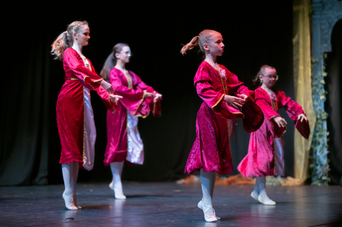 Irene K. - Vier junge Balletttänzerinnen in einheitlichen roten Kostümen und weißen Strumpfhosen treten auf der Bühne auf, halten rote Requisiten in den Händen und nehmen vor einem dunklen Hintergrund eine anmutige Pose ein – dabei zeigen sie, was sie in den örtlichen Tanzkursen gelernt haben.