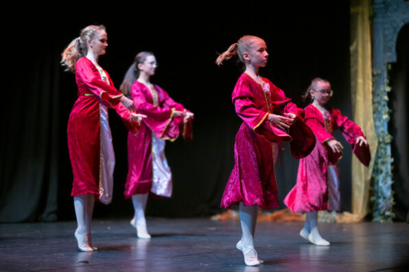 Irene K. - Vier junge Balletttänzerinnen in einheitlichen roten Kostümen und weißen Strumpfhosen treten auf der Bühne auf, halten rote Requisiten in den Händen und nehmen vor einem dunklen Hintergrund eine anmutige Pose ein – dabei zeigen sie, was sie in den örtlichen Tanzkursen gelernt haben.