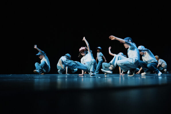 Irene K. - Eine Gruppe von Kindern in Freizeitkleidung und Mützen führt auf einer dunklen Bühne eine Hip-Hop-Tanzchoreografie vor und nimmt dabei dynamische Posen ein, die sie in Tanzkursen gelernt haben – mit erhobenen Armen und angewinkelten Knien.