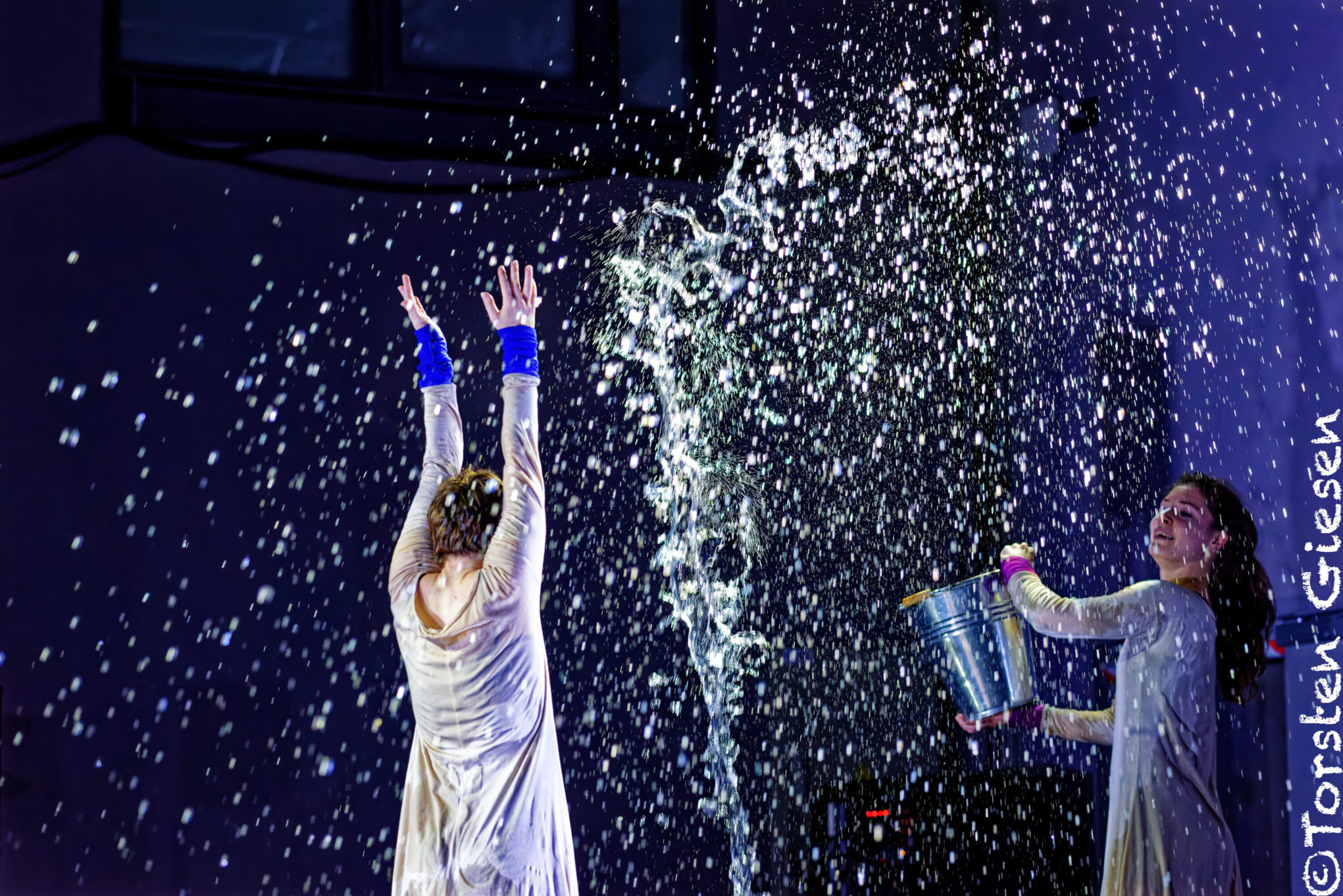 Irene K. - Zwei Performer auf der Bühne führen einen zeitgenössischen Tanz auf, indem sie mit Wasser interagieren. Der eine wirft es aus einem Eimer, während der andere mit erhobenen Armen inmitten dramatischer Spritzer steht. Beide tragen helle Kostüme. Bildnachweis auf der rechten Seite.
