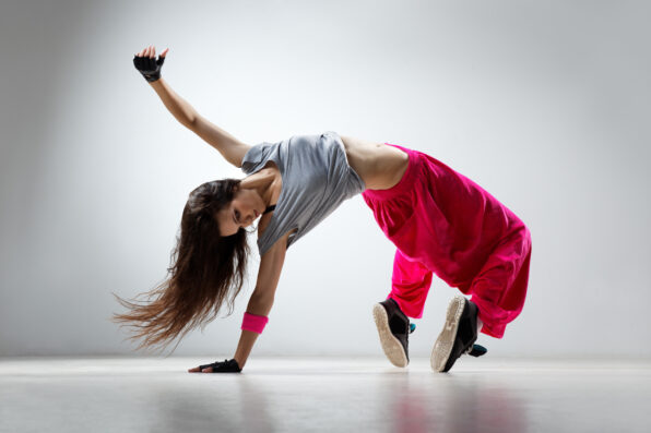 Irene K. - Eine Frau in einem grauen Hemd und einer leuchtend rosafarbenen Hose vollführt eine dynamische Breakdance-Bewegung, wobei sie auf einer Hand balanciert, die Beine ausstreckt und das Haar fließen lässt, vor einem schlichten hellen Hintergrund.