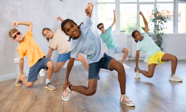 Irene K. - Fünf Kinder in legerer Kleidung nehmen in einem hellen Raum mit Holzfußboden und großen Fenstern energiegeladene Hip-Hop-Tanzposen ein. Sie wirken glücklich und ausdrucksstark, während sie an einer lebhaften Gruppentanzaktivität teilnehmen.