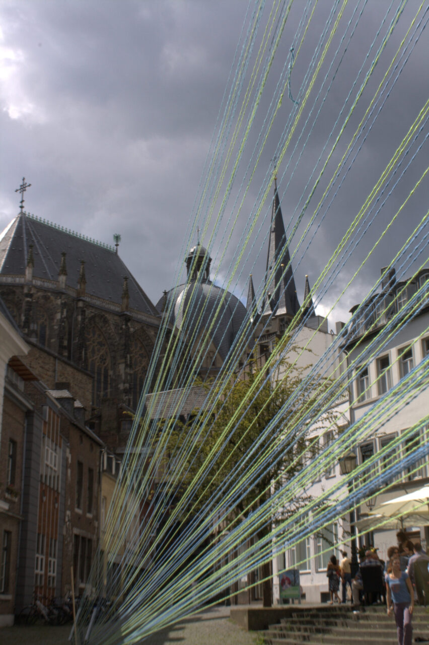 Irene K. - Eine Straßenszene zeigt bunte Schnüre, die diagonal über den Rahmen gespannt sind, mit historischen Gebäuden, einer großen Kirche und Menschen, die unter einem wolkenverhangenen Himmel spazieren gehen - ein Hinweis auf eine ortsspezifische Performance oder einen zeitgenössischen Tanz im öffentlichen Raum.