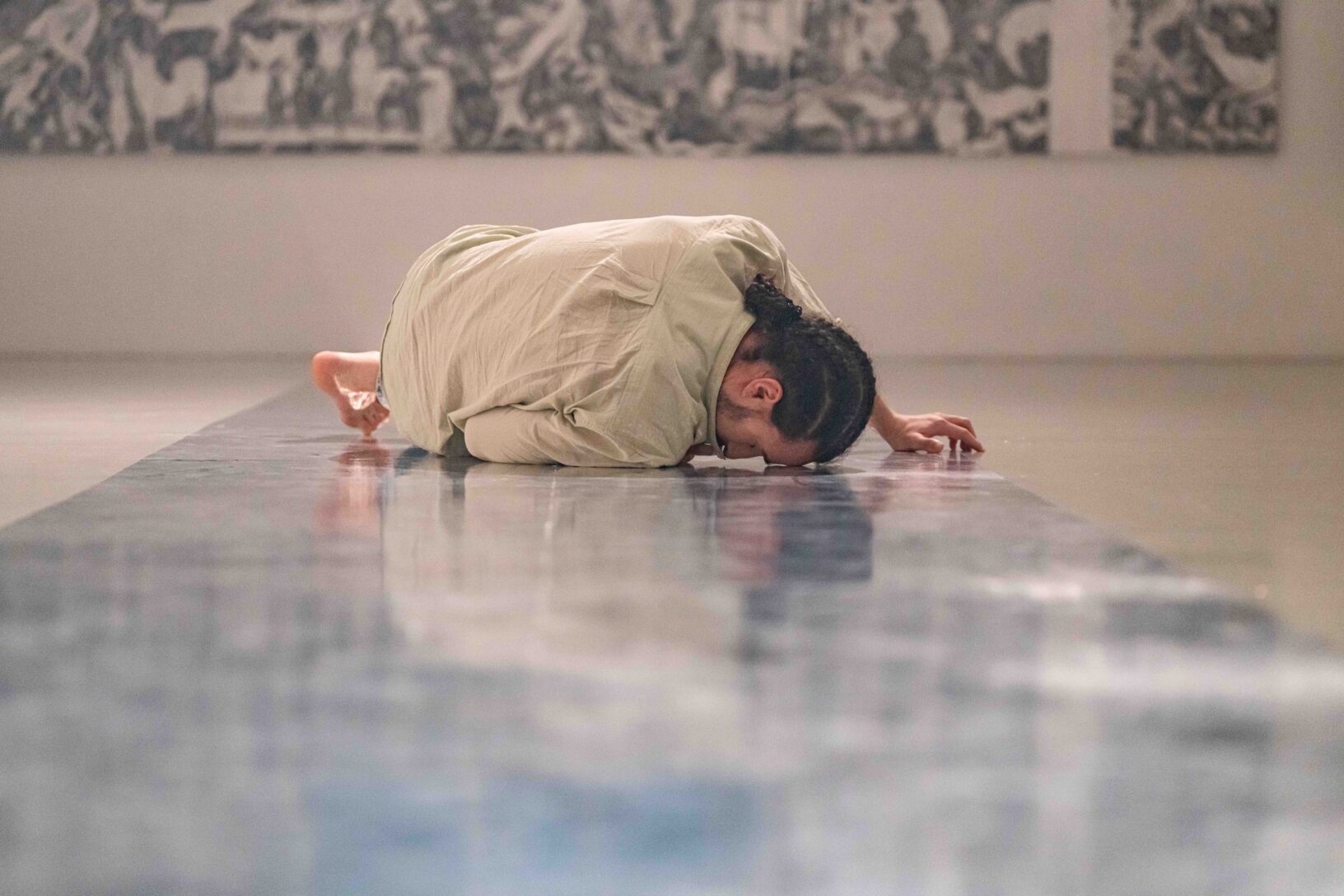 Irene K. - Eine Person in heller Kleidung liegt auf der Seite gerollt auf einem glänzenden Boden und verkörpert zeitgenössischen Tanz in einem minimalistischen, sanft beleuchteten Raum mit abstrakter Kunst an den Wänden - eine stille Begegnung zwischen Bewegung und Raum.