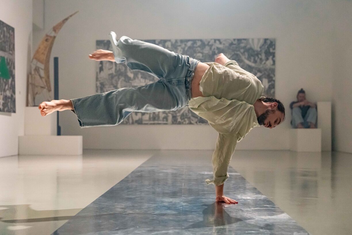Irene K. - Ein Tänzer in lockerem Hemd und Jeans balanciert auf einer Hand, die Beine in der Luft ausgestreckt, in einer Kunstgalerie einen Breakdance-Move - eine dynamische Begegnung von Streetstyle und zeitgenössischem Tanz. Eine sitzende Person schaut aus dem Hintergrund zu.