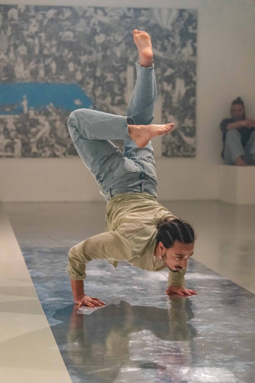Irene K. - Eine Person mit geflochtenen Haaren in Jeans und hellem Hemd balanciert auf den Händen und mit angewinkelten Beinen einen zeitgenössischen Tanz auf einem spiegelnden Boden, hinter dem Kunst an der Wand zu sehen ist.