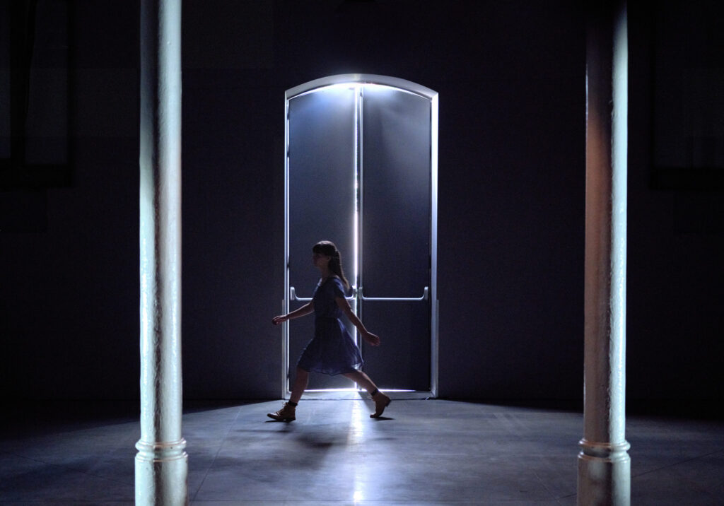 Irene K. - Eine Person in einem Kleid geht durch einen schwach beleuchteten Raum, dessen Silhouette sich vom Licht abhebt, das durch eine offene Tür hinter ihr einfällt, mit zwei weißen Säulen im Vordergrund - eine Szene, die an eine Tanzperformance der Compagnie Irene K. erinnert.