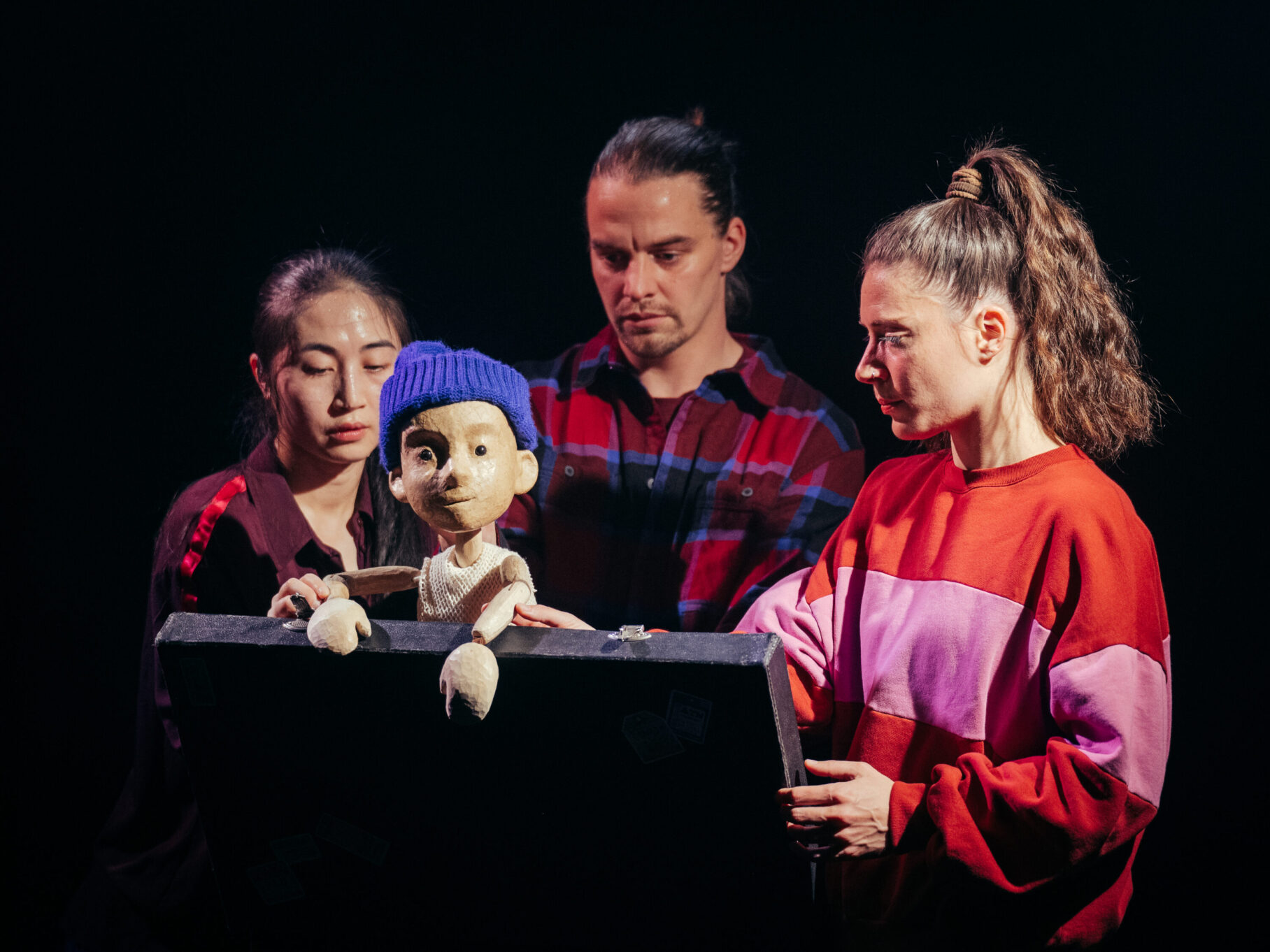 Irene K. - Drei Personen bedienen vorsichtig eine Puppe, die ein Kind mit einer blauen Strickmütze darstellt, das hinter einer schwarzen Fläche in einer dunklen Umgebung steht und sich auf die Aufführung von Tanztheater zum Thema Migration konzentriert.