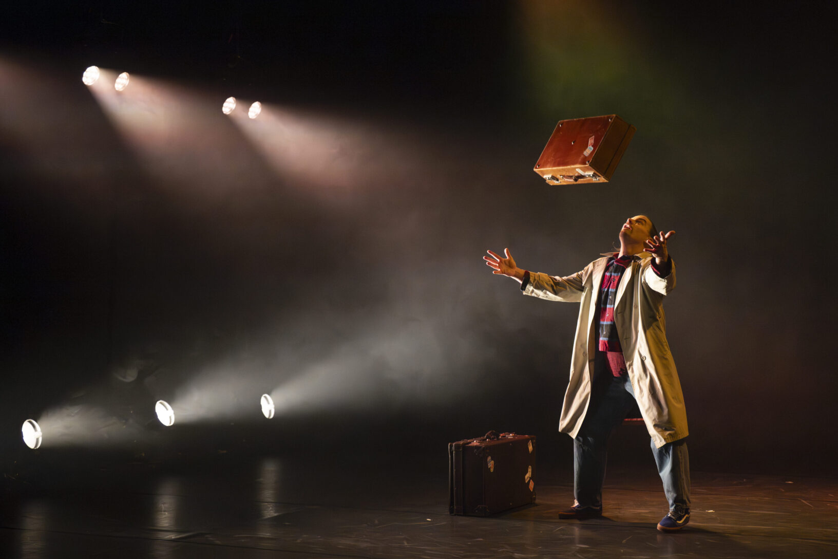 Irene K. - A person in a trench coat and scarf stands on a dimly lit stage, arms outstretched, gazing up at a suitcase tossed in the air. In this evocative Tanztheater by Compagnie Irene K, spotlights shine as another suitcase lies mitgebracht on the floor nearby.