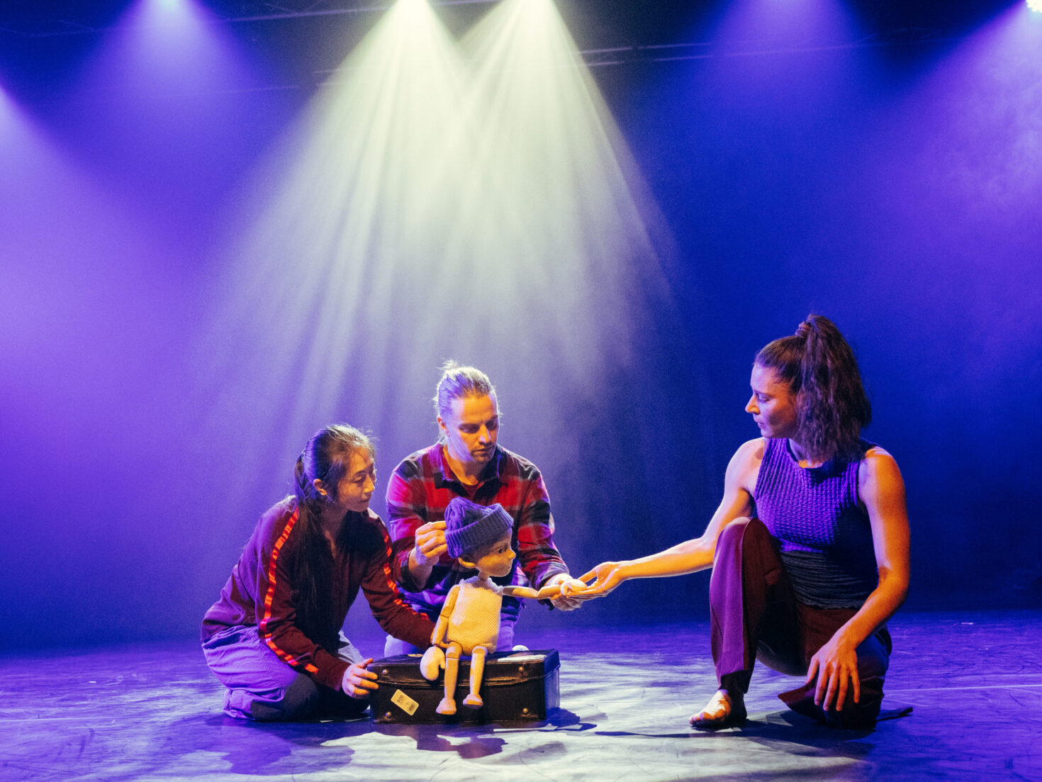 Irene K. - Drei Darsteller auf der Bühne führen eine kleine Puppe in Hut und Pullover, wobei sie Figurenspiel mit eindrucksvoller blauer und violetter Beleuchtung verbinden, während Scheinwerfer in diesem fesselnden Tanztheater für junges Publikum herabstrahlen.