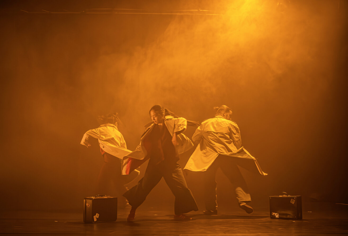 Irene K. - Three dancers from Compagnie Irene K perform Tanztheater on stage, wearing loose jackets amid dramatic orange lighting and fog, two suitcases on the floor beside them—evoking themes of migration.