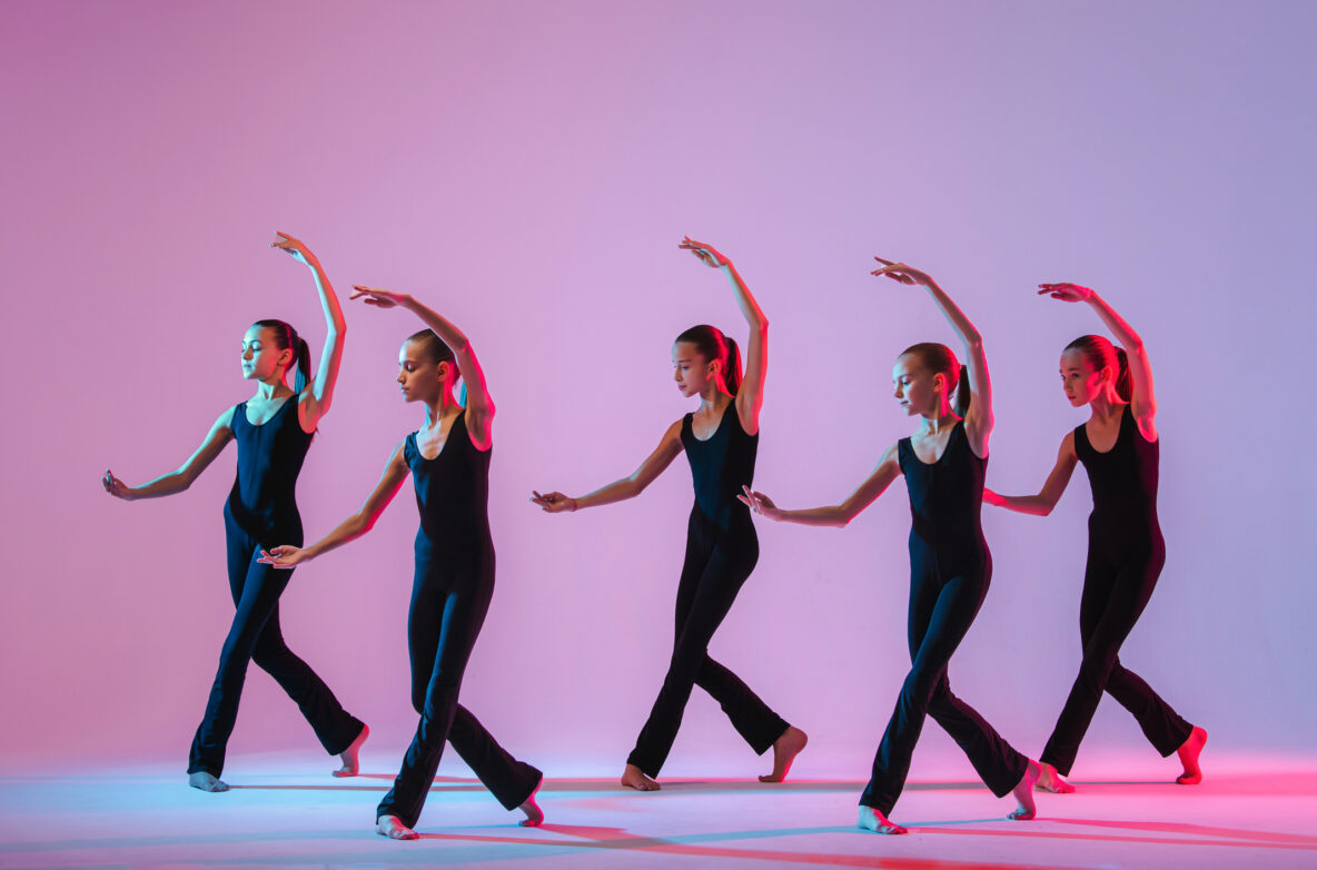Irene K. - Fünf Balletttänzerinnen in schwarzen Trikots führen in einem hell erleuchteten Tanzschulstudio mit rosa und blauer Beleuchtung synchronisierte Bewegungen aus, wobei jede einen Arm anmutig über den Kopf hebt und ein Bein hinter sich ausstreckt.