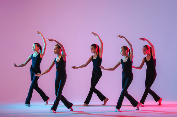 Irene K. - Fünf Balletttänzerinnen in schwarzen Trikots führen in einem hell erleuchteten Tanzschulstudio mit rosa und blauer Beleuchtung synchronisierte Bewegungen aus, wobei jede einen Arm anmutig über den Kopf hebt und ein Bein hinter sich ausstreckt.