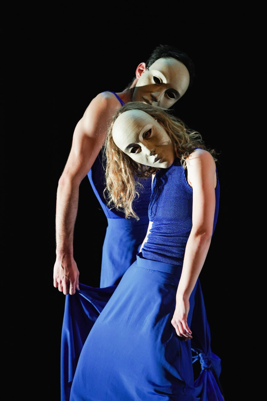 Irene K. - Zwei Tänzerinnen der Compagnie Irene K in blauer Kleidung und weißen Masken posieren eng beieinander vor einem schwarzen Hintergrund und erforschen das Thema Gender Performance, während sie in die Kamera blicken.