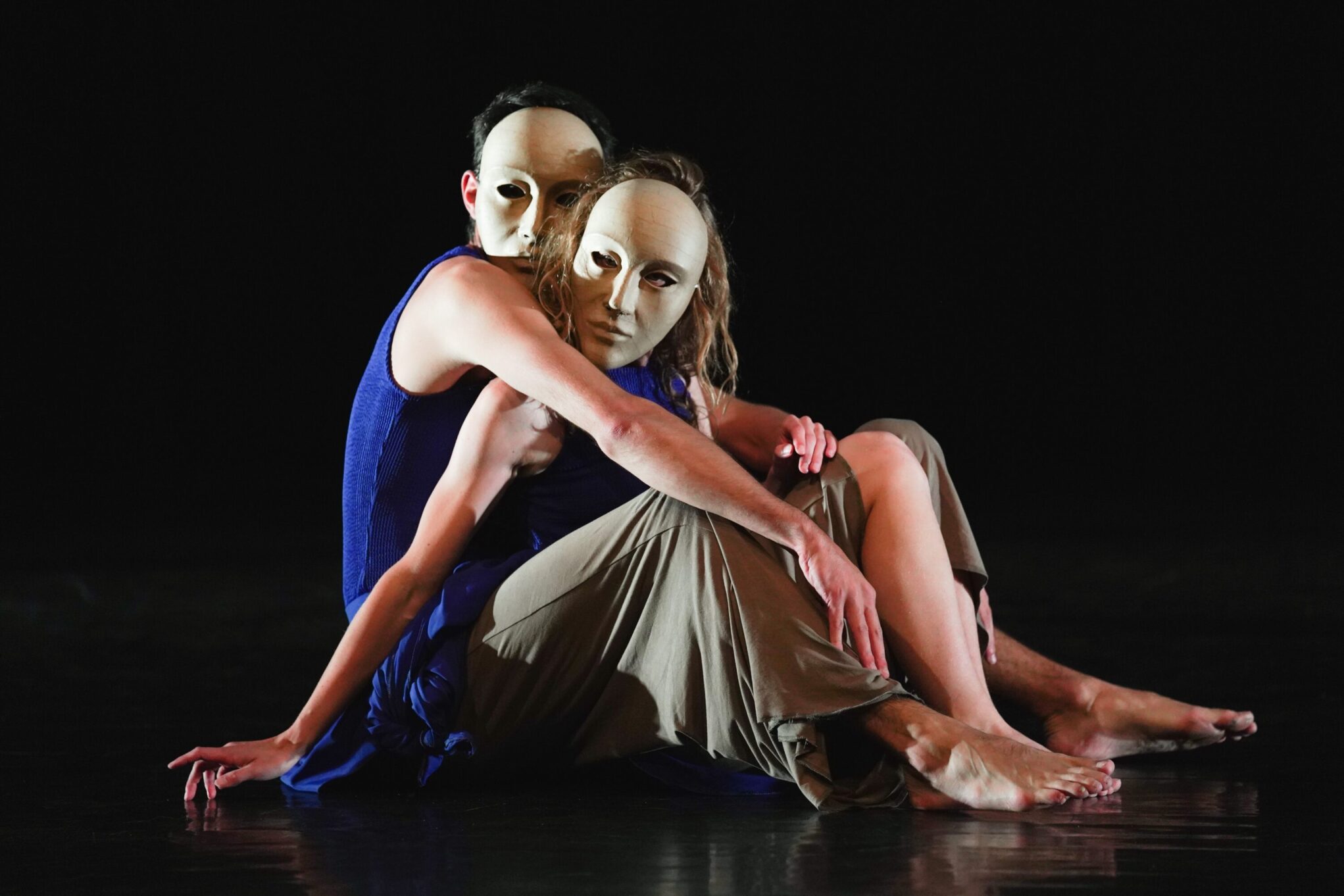 Irene K. - Zwei Tänzer erforschen in einem Tanzduett die Gender-Performance. Sie tragen ausdruckslose weiße Masken und sitzen dicht nebeneinander auf einer dunklen Bühne. In lockerer, fließender Kleidung schaffen sie eine intime Pose, die Identität und Verbindung hinterfragt.