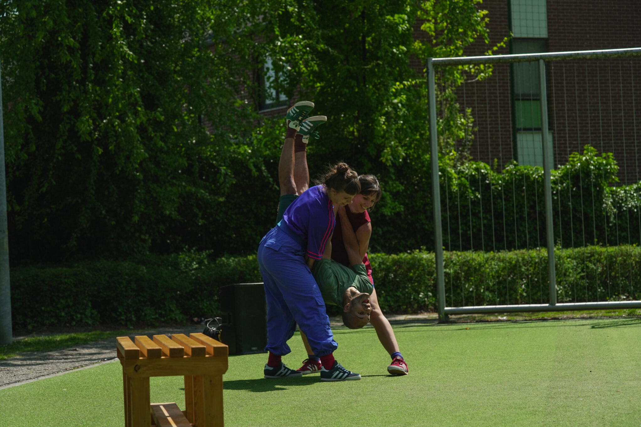 Irene K. - Zwei Personen in farbenfroher Sportkleidung führen während einer Outdoor-Performance der Compagnie Irene K eine akrobatische Bewegung im Freien aus; die eine hält die andere kopfüber an der Taille, die Beine zeigen nach oben, neben einer Holzbank im Gras.