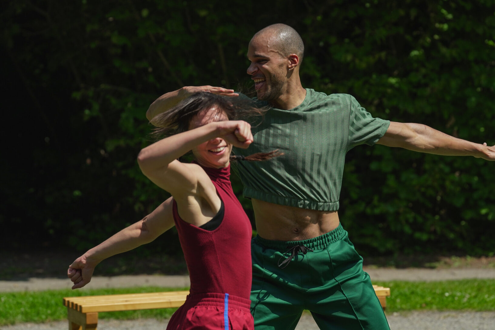 Irene K. - Two people dance energetically outdoors in a lively Outdoor-Performance, smiling with arms outstretched. They wear casual athletic clothes—one in red, the other in green. A wooden bench and green foliage are visible in the background.
