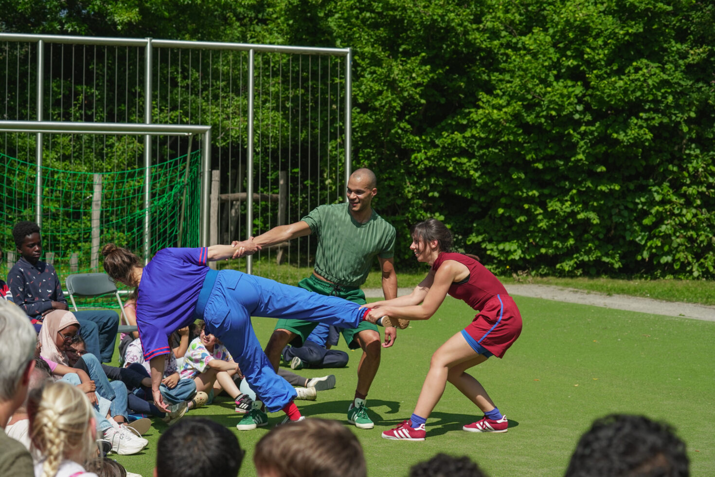 Irene K. - Drei Personen vollführen auf einem grasbewachsenen Sportplatz ein spielerisches Kunststück und verkörpern damit zeitgenössischen Tanz. Einer wird von den beiden anderen waagerecht gehalten, während eine Gruppe von Kindern im Vordergrund diesen lebhaften Tanz im öffentlichen Raum genießt.