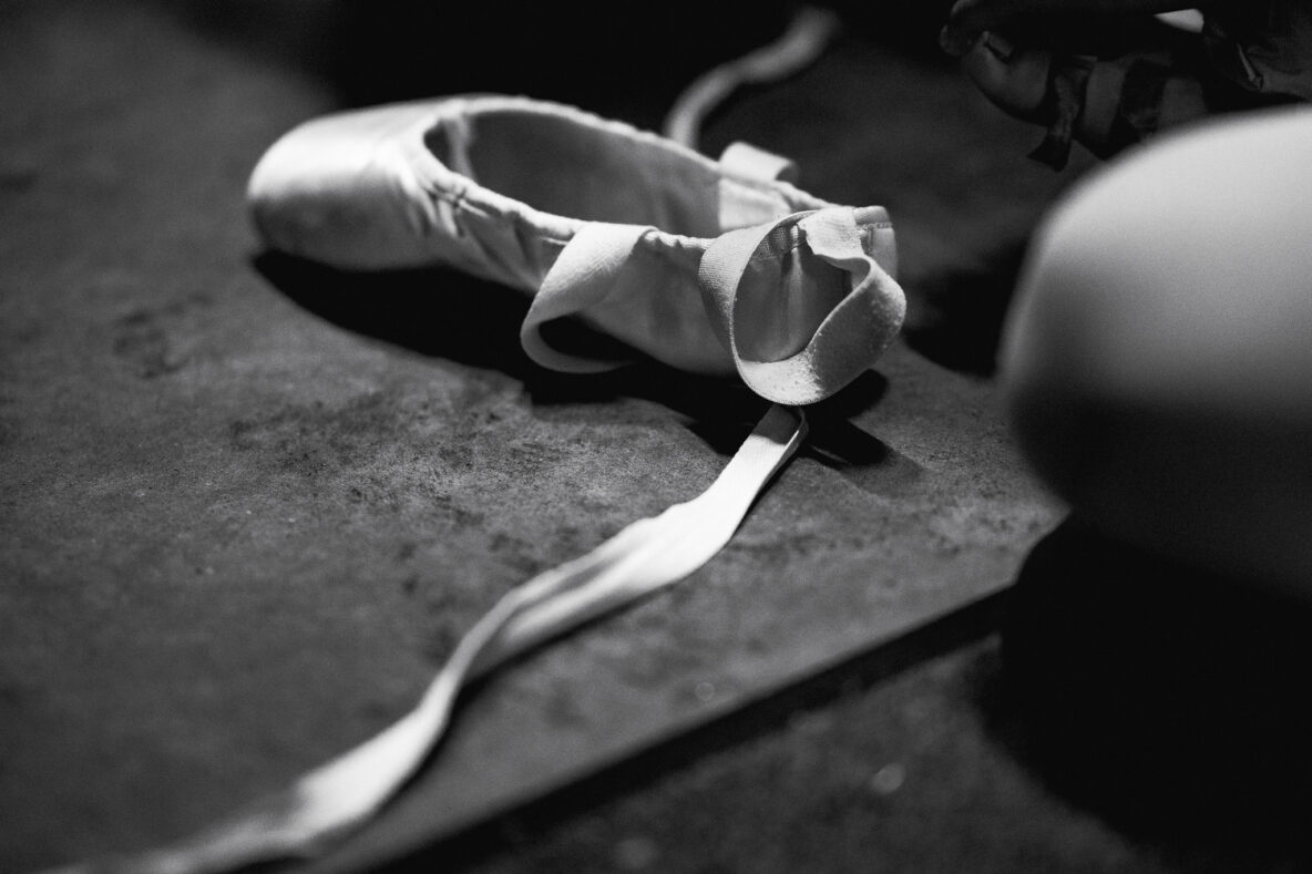 Irene K. - Eine Schwarz-Weiß-Nahaufnahme eines einzelnen Ballettschuhs mit offenem Band, der auf einem strukturierten Boden in einem Studio für Tanzkurse in Verviers liegt. Eine verschwommene Figur ist teilweise auf der rechten Seite des Bildes zu sehen.