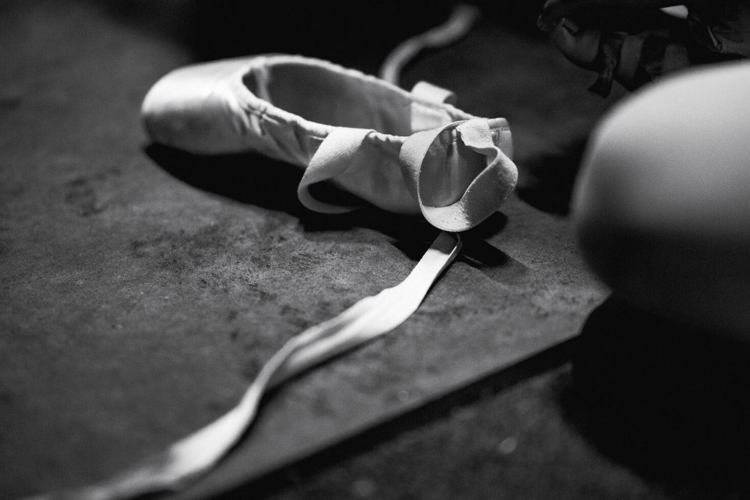 Irene K. - Eine Schwarz-Weiß-Nahaufnahme eines einzelnen Ballettschuhs mit offenem Band, der auf einem strukturierten Boden in einem Studio für Tanzkurse in Verviers liegt. Eine verschwommene Figur ist teilweise auf der rechten Seite des Bildes zu sehen.