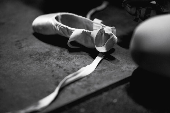Irene K. - Eine Schwarz-Weiß-Nahaufnahme eines einzelnen Ballettschuhs mit offenem Band, der auf einem strukturierten Boden in einem Studio für Tanzkurse in Verviers liegt. Eine verschwommene Figur ist teilweise auf der rechten Seite des Bildes zu sehen.
