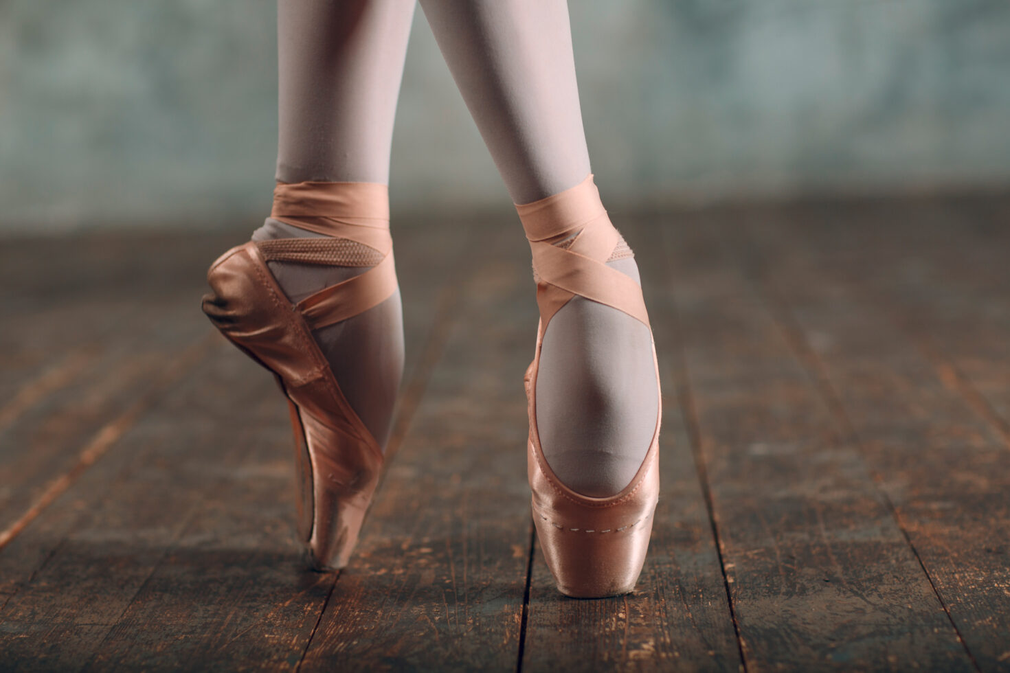 Irene K. - Nahaufnahme der Füße einer Balletttänzerin in rosafarbenen Spitzenschuhen, die auf einem abgenutzten Holzboden steht, die Beine gekreuzt und die Knöchel mit Bändern umwickelt. Der verschwommene Hintergrund deutet auf eine Tanzschule hin, in der sich Modern Jazz und klassische Stile vermischen.