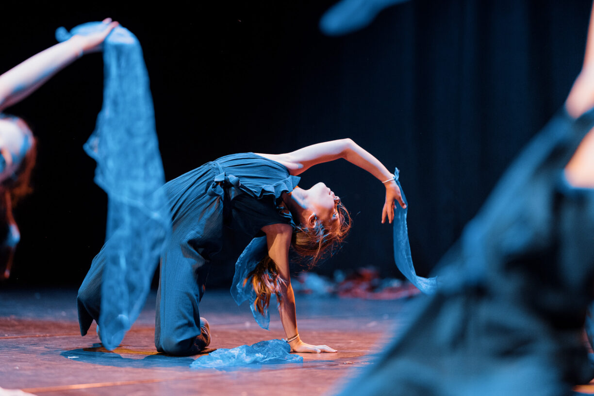 Irene K. - A dancer in a dark jumpsuit kneels on stage, arching backward with one arm extended and holding a blue scarf—a striking moment of zeitgenössischer Tanz. In the shadowy background, other dancers with scarves are partially visible.