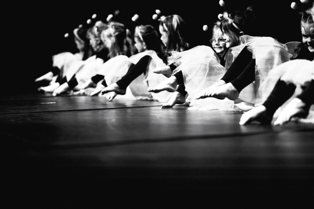 Irene K. - A row of young children in costumes sits on stage with their feet extended forward in a dance pose; the black and white image with dramatic lighting captures their joyful expressions and matching outfits from a lively Hip-Hop Tanzkurse performance.