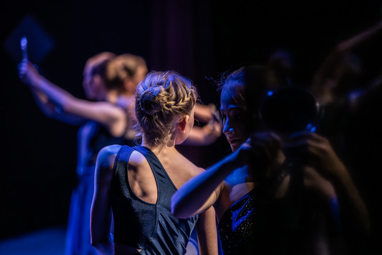 Irene K. - Zwei Frauen in eleganten Kleidern stehen auf einer schwach beleuchteten Bühne in einer Tanzschule, eine davon mit geflochtenem blondem Haar, die den Blick abwendet. Andere im Hintergrund halten Spiegel und schaffen eine dramatische und stimmungsvolle Aufführungsszene mit tiefblauer und violetter Beleuchtung.
