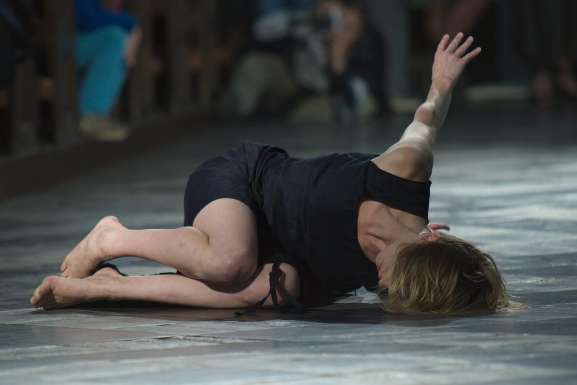 Irene K. - Eine barfüßige Tänzerin in einem schwarzen Kleid liegt auf dem Boden, den Arm ausgestreckt, den Kopf nach unten geneigt, während einer dramatischen zeitgenössischen Tanzaufführung.