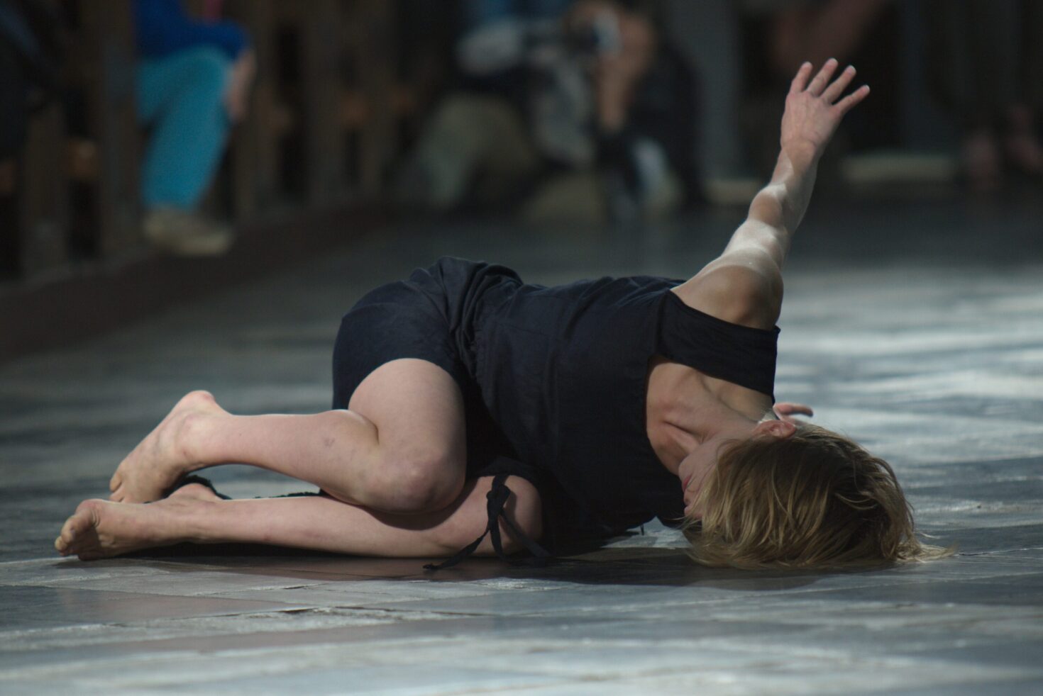 Irene K. - Eine barfüßige Tänzerin in einem schwarzen Kleid liegt auf dem Boden, den Arm ausgestreckt, den Kopf nach unten geneigt, während einer dramatischen zeitgenössischen Tanzaufführung.